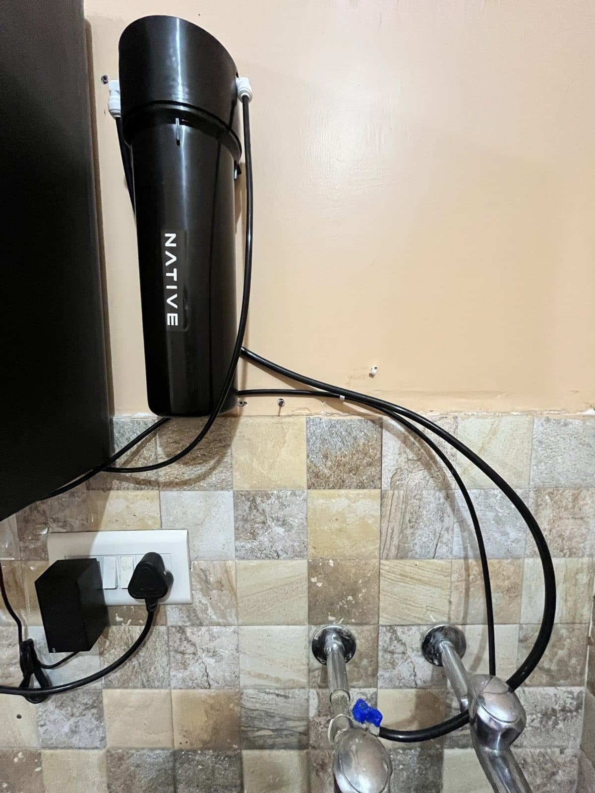 The black Native pre-filter canister fixed to the wall beside the purifier, with the inlet pipe running up from a blue-handled valve on the kitchen tap line.