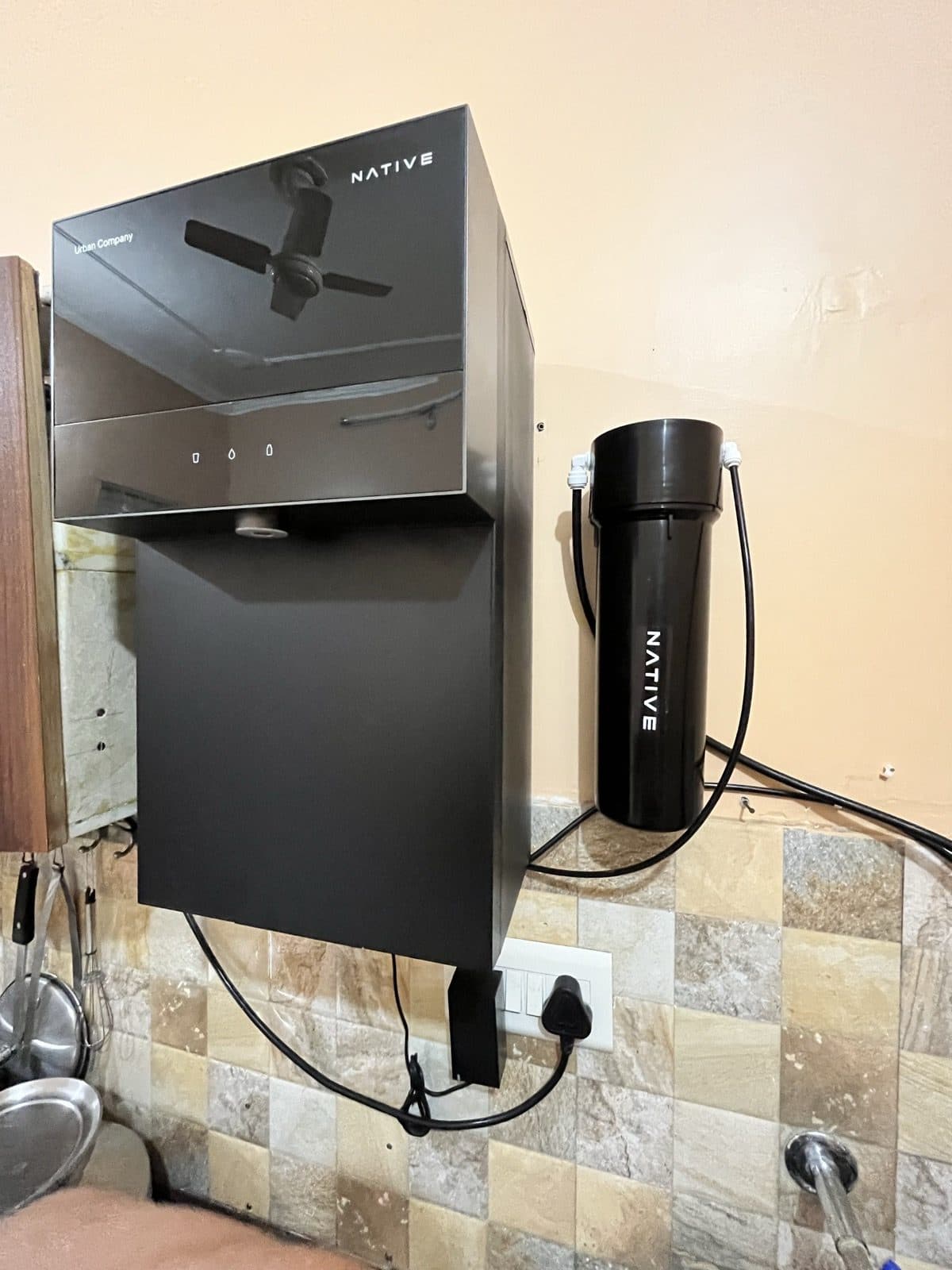 The Native M3 Pro mounted on a tiled kitchen wall, seen at an angle, with the black pre-filter canister beside it and pipes looping between them.